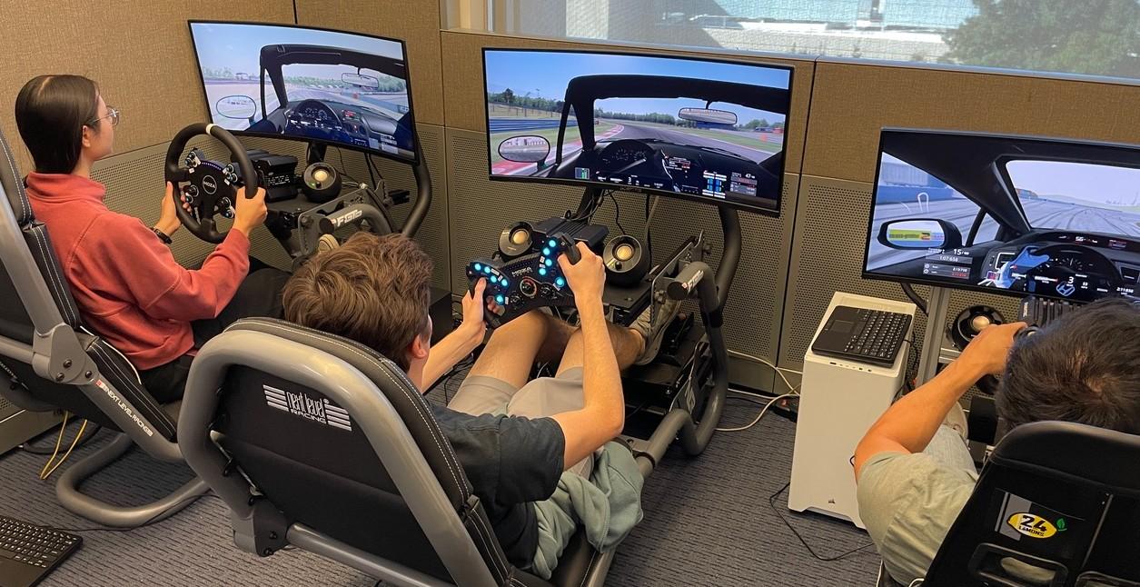 Three students, one in an orange sweater, one in a short sleeve black shirt and one in beige shirt, practice high speed driving on realistic video game simulators. 