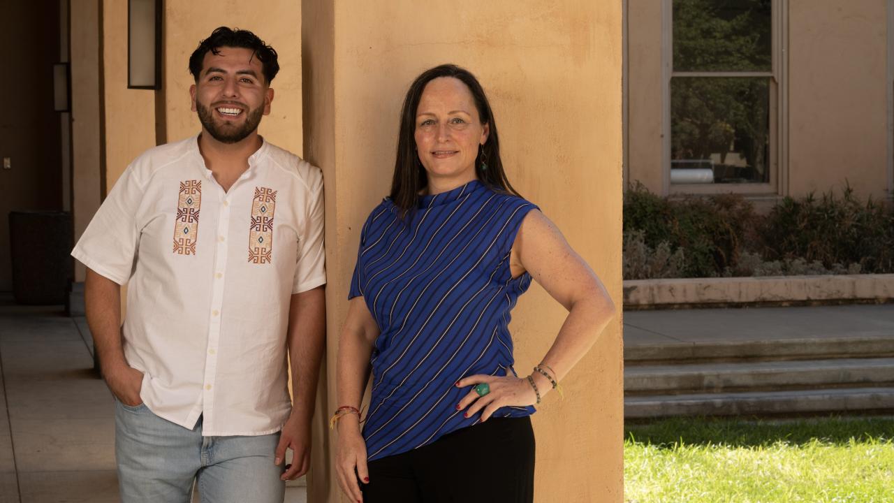 A man and a woman stand smiling side by side outside a tan stucco building. The man is wearing a white embroidered short-sleeve shirt and jeans, while the woman is wearing a blue and black striped sleeveless top and black pants. They are posed casually on either side of a square column, with sunlight casting soft shadows on the scene. A grassy lawn and window are visible in the background.