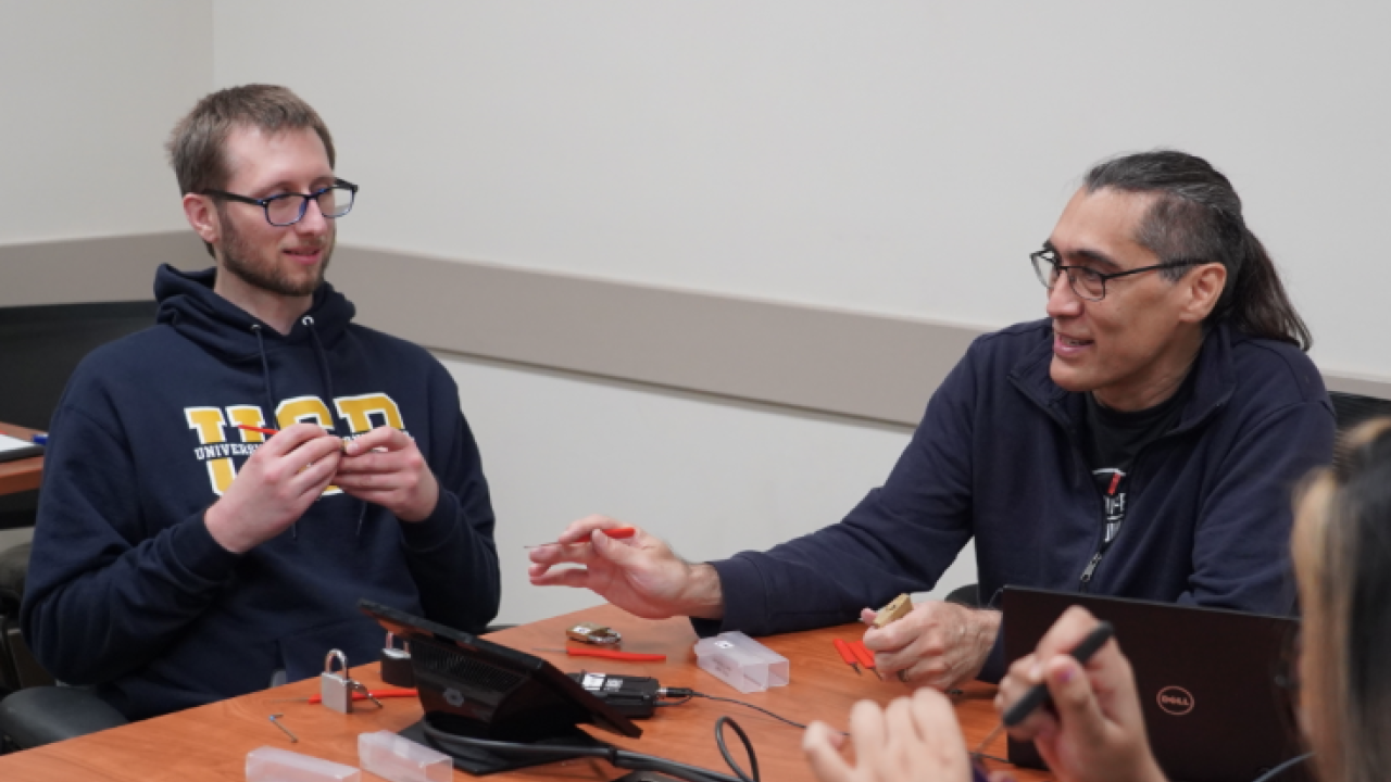 A professor talks to a student as they practice how to pick a lock. They are seated in a UC Davis classroom around a table full of locks and small tools. 